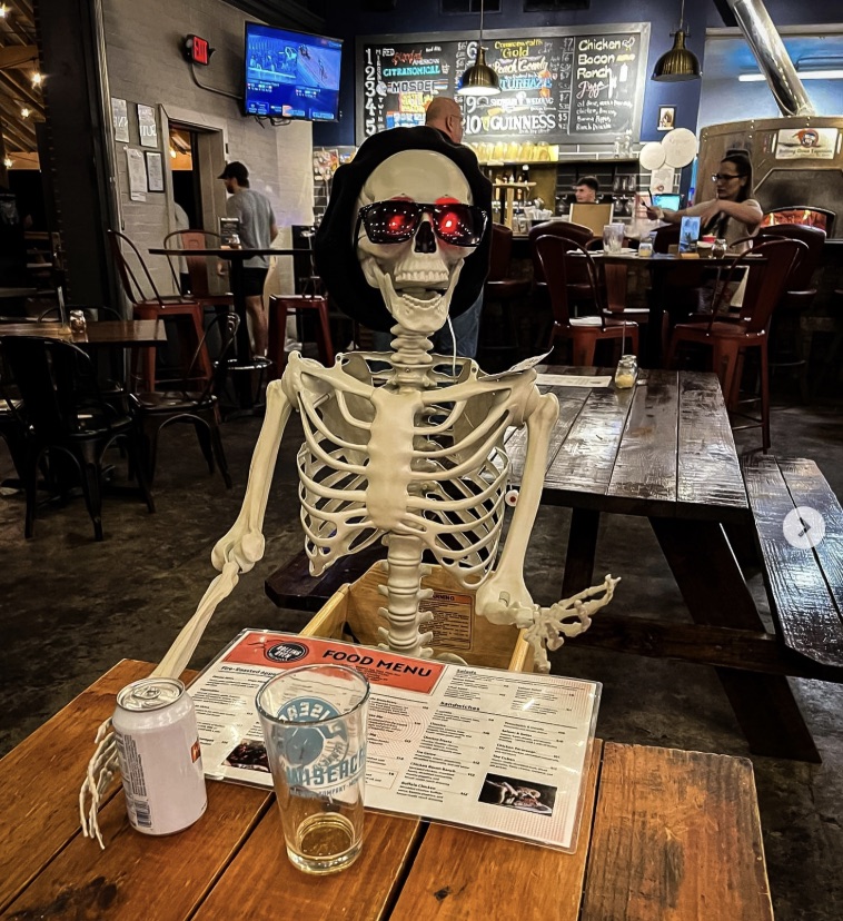 A plastic skeleton in sunglasses and a beanie, sitting at a bar table with a menu and a beer